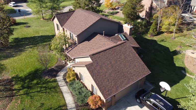 An aerial view of a two-story suburban house with a brown roof, landscaped lawn, and a car parked in the driveway.