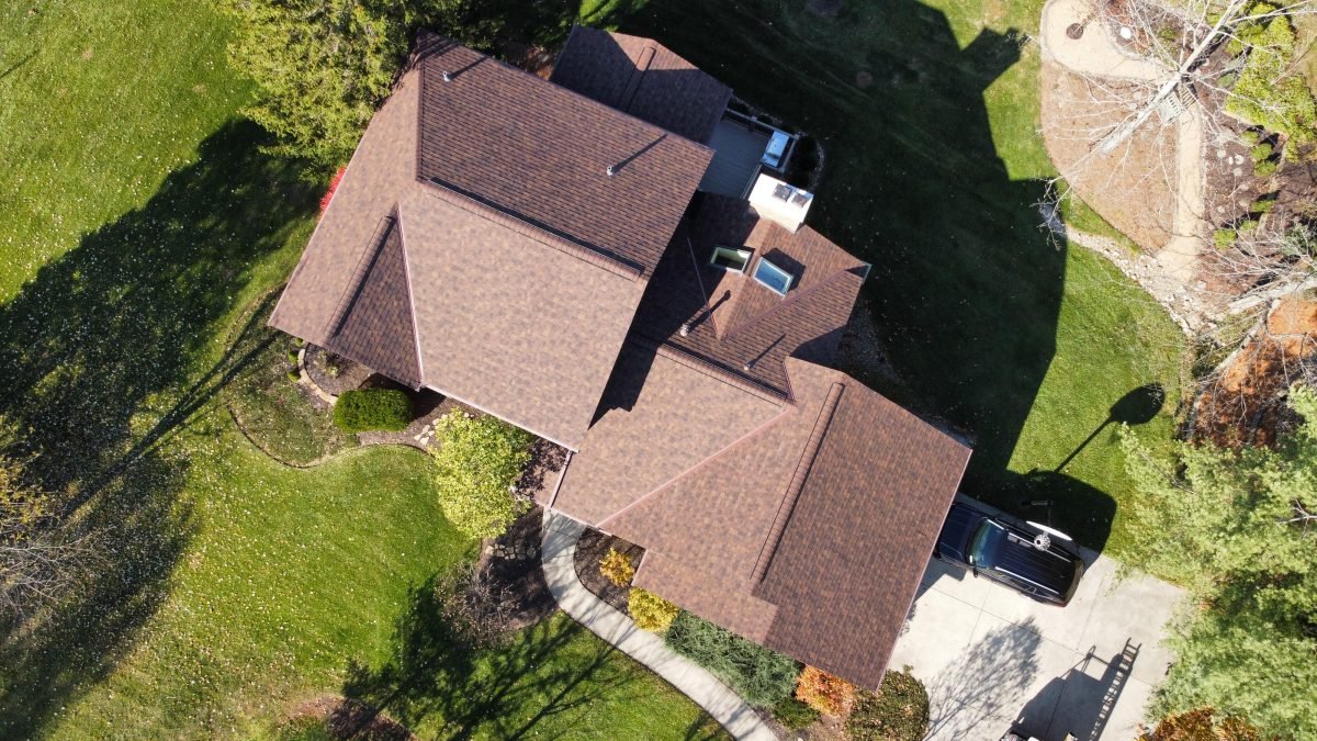 An aerial view of a large house by a lumbus Ohio Roofing Contractor with a brown roof surrounded by trees and a lawn with a driveway on the side. -residential roofing services in Central Ohio