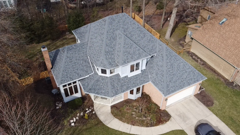 Aerial view of a two-story residential house with gray shingles, a driveway, and surrounding trees.