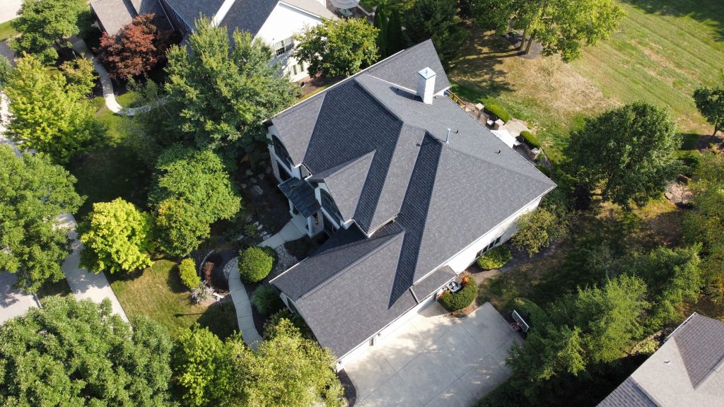 An aerial view of a large house with a gray roof, surrounded by a well-manicured lawn and trees, in a suburban neighborhood.