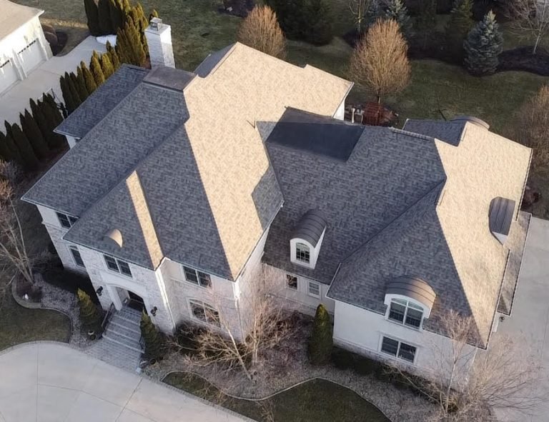 An aerial view of a large two-story house with a gray roof and beige exterior, surrounded by a landscaped yard.
