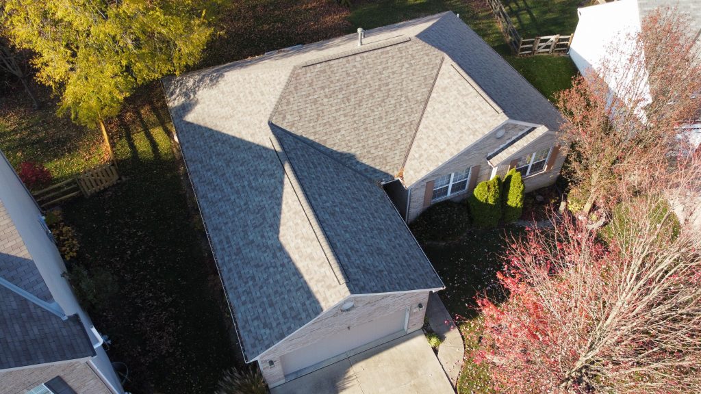 An aerial view of a suburban house with a gray shingle roof, surrounded by a lawn and trees, in the daylight.