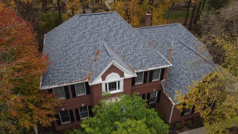 The image shows an aerial view of a two-story brick house with a gray shingle roof surrounded by trees with autumn foliage.