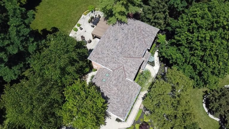 An aerial view of a house with a gray roof surrounded by green trees and a well-manicured lawn with a patio area.