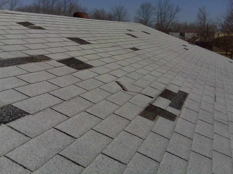 A damaged shingle roof with multiple missing pieces exposing the underlayment, in need of repair.