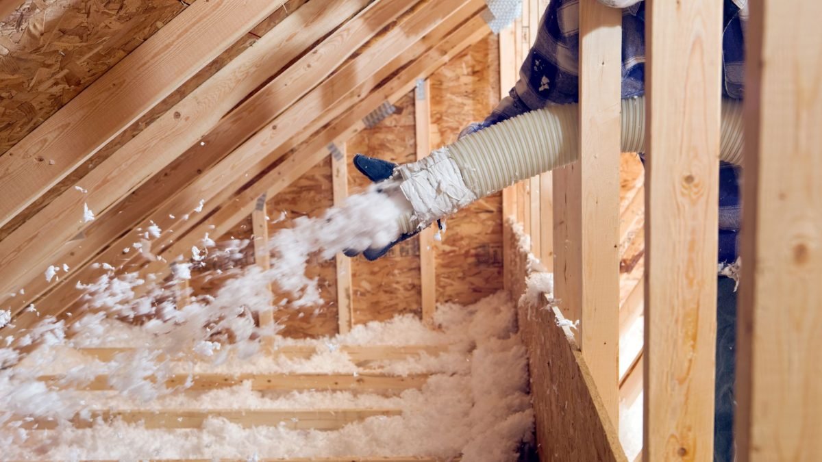 A worker is installing blown-in insulation in the attic of a house between wooden rafters.