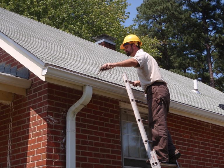 A worker wearing a yellow helmet is using a ladder to reach the roof of a brick house for maintenance or inspection.