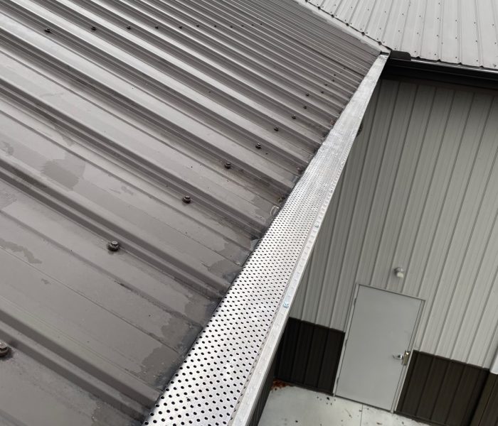 A close-up view of a perforated metal gutter guard installed on a sloping metal roof against a cloudy sky backdrop.